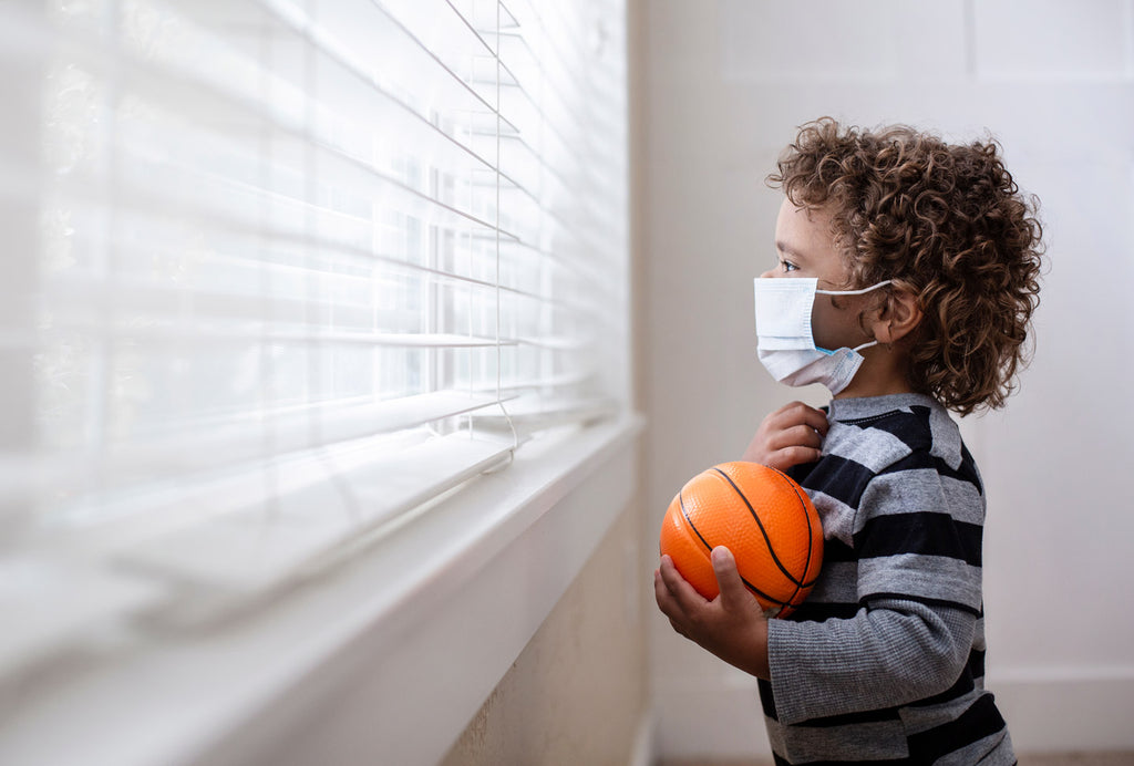 Young boy wearing mask and holding basketball looks out of the window by himself to avoid social interaction.