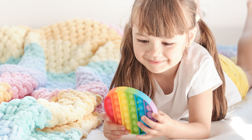 Little cheerful girl plays with a rainbow colored plastic pop fidget toy while lying on the bed, a useful toy that may help with perservation in autism.